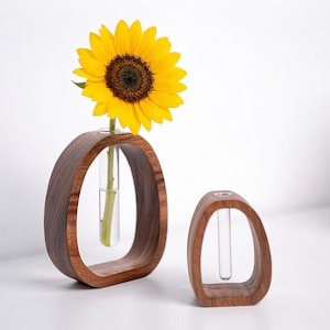 Op de afbeelding: Twee houten bloemknoppen, een met een heldergele zonnebloem. De vazen hebben een donkerbruine afwerking en een ovale vorm met een heldere glazen buis voor het vasthouden van een enkele stengel. De zonnebloem heeft een donkerbruin hart en gele bloemblaadjes.