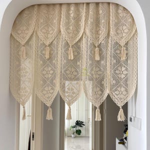 Puede incluir: Cortina de puerta de ganchillo color crema. La cortina presenta un patrón floral repetitivo y está adornada con borlas en la parte inferior de cada panel. La cortina cuelga en una puerta arqueada, agregando un toque de encanto vintage al espacio.