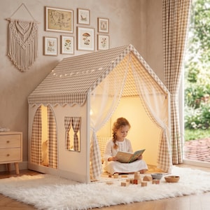 以下が含まれることがあります： ベージュと白のチェック柄の屋根とカーテンが付いた子供用のプレイハウスのテント。 テントには窓があり、ふわふわの白いラグの上に設置されています。 少女が中に座って本を読んでいます。 装飾的な額入りのアートが壁に掛けられています。