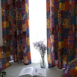 Puede incluir: Cortinas estampadas con patchwork en tonos naranja, morado y azul. Las cortinas están hechas de varios cuadrados de tela con diseños de cachemira y geométricos. Un jarrón con flores secas y un libro abierto están en el alféizar.
