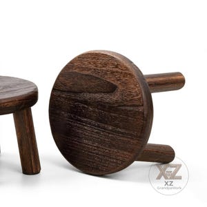 May include: Two small, dark brown wooden stools. One stool is upright, showing the seat and a leg. The other stool is flipped over, revealing the underside of the seat and two legs. The wood grain is visible.