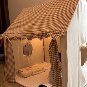 以下が含まれることがあります： 茶色と白のチェック柄の屋根とカーテンが付いた子供用プレイハウスのテント。テントは白で、窓が2つ開いています。内部にはマットレスと枕があります。扇風機が見えます。