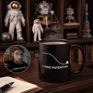 May include: A black ceramic mug with the text "I HAD POTENTIAL" in white, with a white line and a small white ball. The mug contains a hot drink, with steam rising. The background features astronaut figurines and a bookshelf.