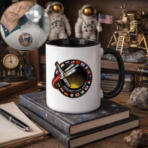 Puede incluir: Taza de cerámica blanca con asa y borde negros. La taza presenta un diseño circular con un cohete, un sol y el texto "PROJECT HAIL MARY" rodeado de banderas. La taza está sobre una pila de libros.