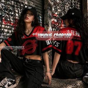 May include: Black and red cropped jersey top with the text "UNIQUE CROPTOP" and "BTS" printed on it. The top has a V-neck and short sleeves. The number "07" is printed on the back. The model is wearing a black baseball cap and black pants.