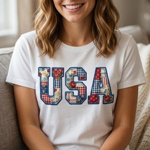 Op de afbeelding: Een wit T-shirt met korte mouwen en de letters USA op de borst. De letters zijn gevuld met een patchworkpatroon van rode, witte en blauwe bloemen- en vichy-ruitstoffen, omlijnd met een dikke blauwe rand voor een opvallende uitstraling.