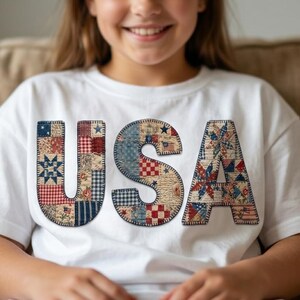 Puede incluir: Una camiseta blanca de manga corta con las letras USA en el pecho. Cada letra está rellena con un patrón de patchwork en tonos rojos, blancos y azules, incluyendo estrellas, rayas y motivos florales con costuras visibles.
