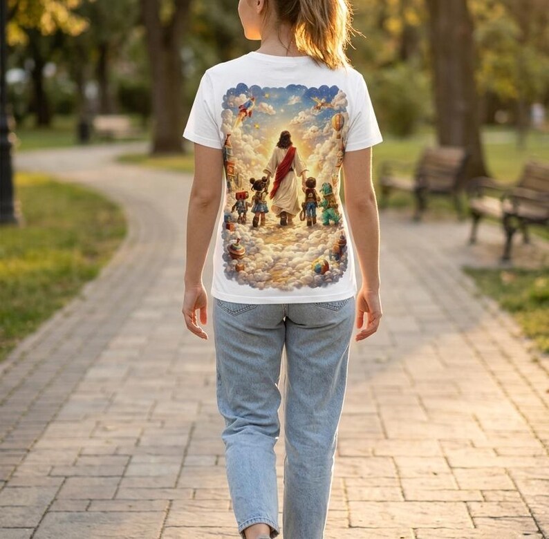 May include: White t-shirt featuring a colorful illustration of a religious figure leading children through clouds. The design includes a blue sky, hot air balloons, and other whimsical elements. The shirt is made of a soft material.