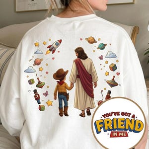 May include: White sweatshirt featuring a colorful graphic of Jesus holding hands with a cowboy, surrounded by planets, stars, and toys. The text "YOU'VE GOT A FRIEND IN ME" is in a circular design on the lower right.