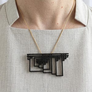 May include: A black geometric pendant necklace with a gold chain. The pendant features a layered, rectangular design with a series of nested shapes. The necklace is worn against a light gray linen top.