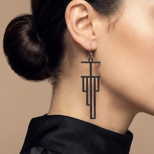 May include: Black geometric dangle earrings. The earrings have a linear design with a horizontal bar at the top and vertical lines extending downwards. The earrings are suspended from a small hook. The earrings are worn by a person with dark hair.
