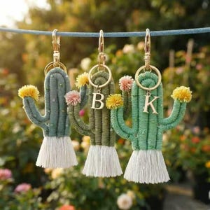 Op de afbeelding: Drie cactusvormige sleutelhangers gemaakt van omwikkeld groen garen, elk met een witte kwast aan de basis en kleine gele of roze pompons op de armen. Twee van de sleutelhangers bevatten goudkleurige metalen letterbedels, namelijk B en K.
