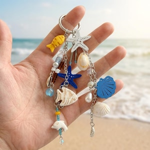 May include: A silver-toned metal keyring featuring multiple dangling charms including a yellow fish, a white starfish, a blue starfish, a white dolphin, a blue seashell, and various beads. The charms are attached to a circular metal ring.