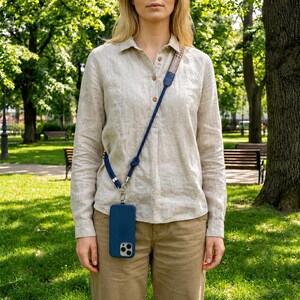 Puede incluir: Una funda de smartphone azul oscuro sujeta a una correa larga y ajustable. La correa presenta una sección tejida con patrones multicolores cerca del hombro y un cordón azul oscuro sólido que se conecta a la funda mediante herrajes de metal plateado.