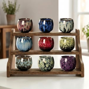 Puede incluir: Una estantería de madera exhibe nueve tazas de té de cerámica en varios colores, incluyendo azul, rojo, verde y morado. Las tazas tienen un esmalte moteado y están dispuestas en tres filas de tres. La estantería es de madera marrón claro.