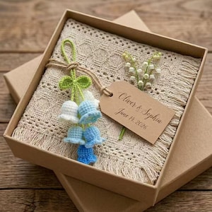 May include: A beige crocheted blanket with fringe is presented in a brown box. A tag reads "Oliver & Sophia June 14, 2026". Crochet flowers in white and blue are attached with twine. A sprig of lily of the valley is also included.