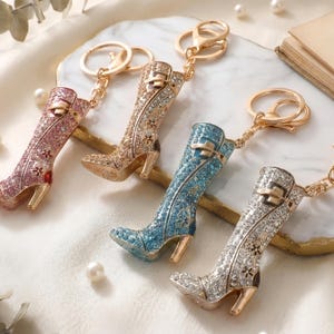 May include: Four rhinestone-studded boot keychains in pink, gold, blue, and silver. Each boot has a gold-colored heel and a matching gold keychain ring. The boots are arranged on a white surface with pearls.