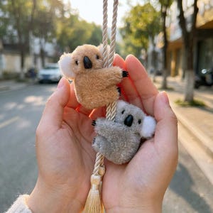 May include: Two plush koala bear ornaments, one tan and one gray, hang from a beige rope with a tassel. The koalas have black noses, small black claws, and white ear tufts. The background is blurred.