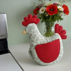 May include: A chicken-shaped oven mitt in shades of grey and red. The mitt features a floral pattern, a red comb, wattle, and tail, and a red patch on the front. A vase of flowers is in the background.