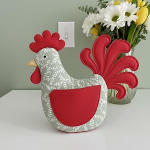 Può includere: Un oggetto decorativo in tessuto a forma di gallo. Il corpo è grigio chiaro con un motivo a foglie bianche, e la cresta, il bargiglio e la coda sono rossi. Ha un becco giallo ed è posto su una superficie bianca.