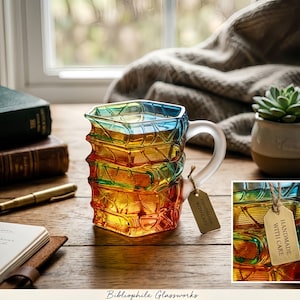 May include: A multi-coloured glass mug with a distinctive stacked book design. The mug transitions from blue to yellow to red, with a clear handle and a small tag that reads "HANDMADE WITH CARE". The mug is on a wooden surface.