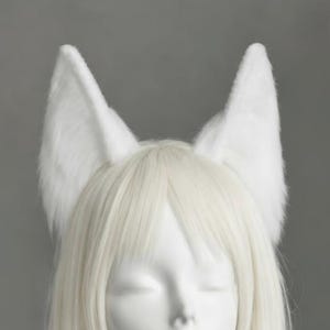 May include: White faux fur fox ears on a headband. The ears are upright and fluffy, with a pointed shape. The ears are positioned on a mannequin head with long, straight, white hair. The background is a solid grey.