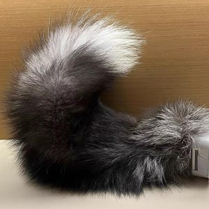 Puede incluir: Una cola de piel sintética en tonos negro, gris y blanco. La cola está unida a una base de plástico blanco con correas de plástico transparente. La cola está rizada y descansa sobre una superficie clara.