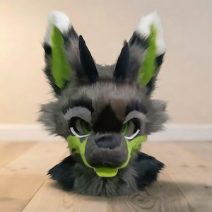 以下が含まれることがあります： グレー、黒、緑色の毛皮のオオカミの頭。耳は白と緑で縁取られ、目は緑色です。マズルと内側の耳は明るい緑色です。コスプレや衣装に最適な、リアルで漫画的なスタイルです。
