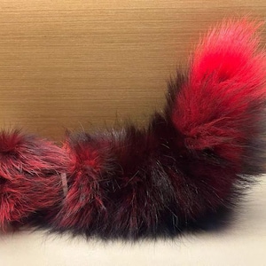 以下が含まれることがあります： 赤と黒のふわふわフェイクファーの尻尾、白いアタッチメント付き。尻尾は黒から赤へと色が変化しています。白い表面に置かれ、背景は茶色です。