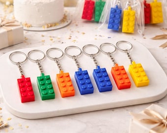 May include: A collection of colorful Lego brick keychains in red, green, orange, blue, and yellow. The keychains are displayed on a white tray, with some packaged in clear bags. A small cake with lit candles is in the background.