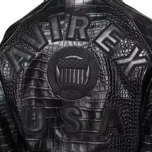 May include: A black jacket with a textured crocodile skin pattern. The back features large black block letters spelling AVIREX and USA. A central circular emblem displays a shield with stars and stripes and the text ESTABLISHED 1975.