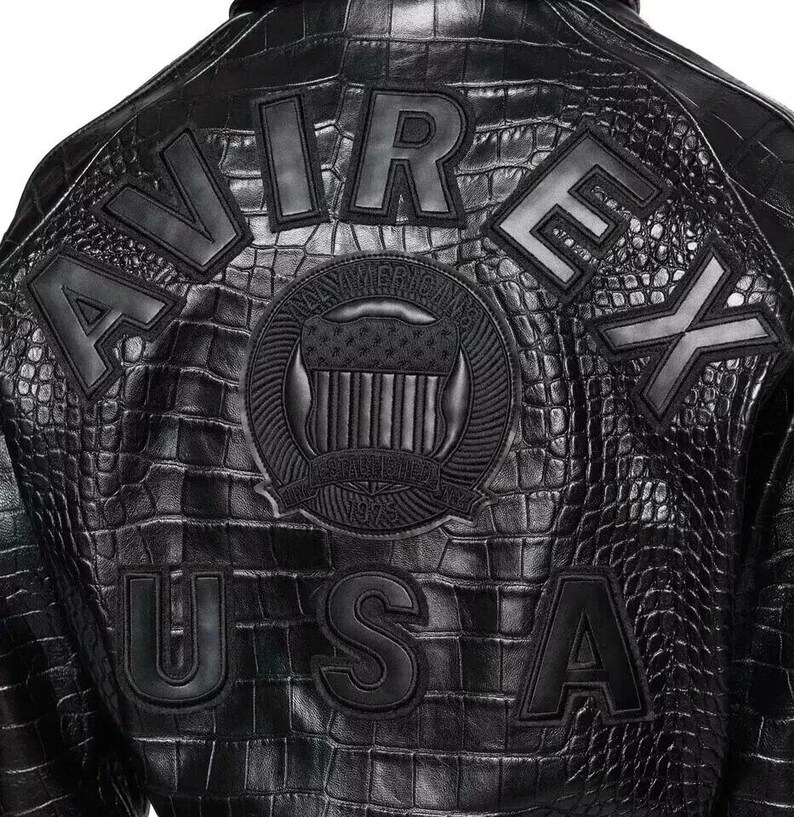 May include: A black jacket with a textured crocodile skin pattern. The back features large black block letters spelling AVIREX and USA. A central circular emblem displays a shield with stars and stripes and the text ESTABLISHED 1975.