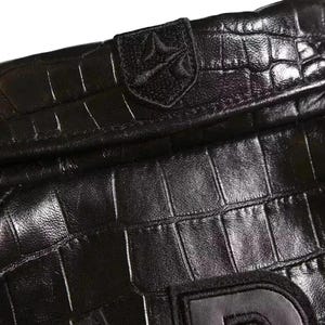 May include: A black leather item with a textured crocodile skin pattern. The surface features a stitched emblem with a shield-like shape containing a stylized symbol. A portion of a large, dark letter is visible at the bottom edge of the frame.