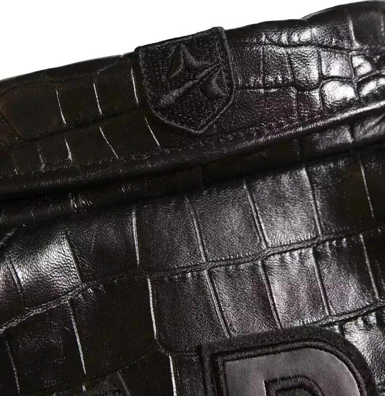 May include: A black leather item with a textured crocodile skin pattern. The surface features a stitched emblem with a shield-like shape containing a stylized symbol. A portion of a large, dark letter is visible at the bottom edge of the frame.
