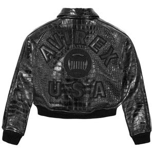 May include: A black jacket with a textured crocodile-skin pattern. The back features the embossed text AVIREX USA in a semi-circular arrangement above a circular logo. The garment includes a collar, long sleeves, and a black ribbed knit hem.