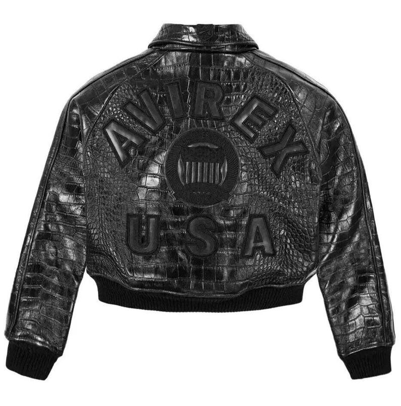 May include: A black jacket with a textured crocodile-skin pattern. The back features the embossed text AVIREX USA in a semi-circular arrangement above a circular logo. The garment includes a collar, long sleeves, and a black ribbed knit hem.