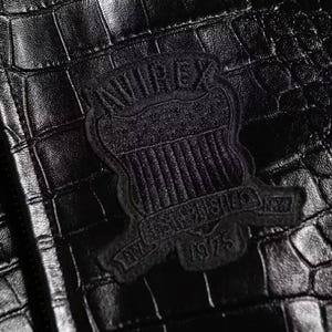 May include: A black jacket with a textured crocodile skin pattern. Centered on the material is a black embroidered patch featuring the text AVIREX, ESTABLISHED, and 1975, arranged in a shield-like emblem design with vertical stripes.