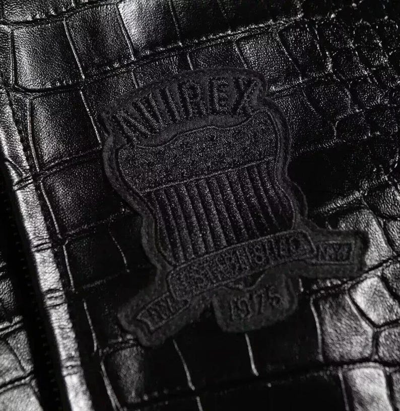 May include: A black jacket with a textured crocodile skin pattern. Centered on the material is a black embroidered patch featuring the text AVIREX, ESTABLISHED, and 1975, arranged in a shield-like emblem design with vertical stripes.