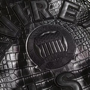 May include: A black material with a crocodile skin texture featuring large black stitched lettering and a circular emblem. The emblem displays a shield with stars and stripes, the text AMERICANS, ESTABLISHED, 1979, and NY in a circular border.