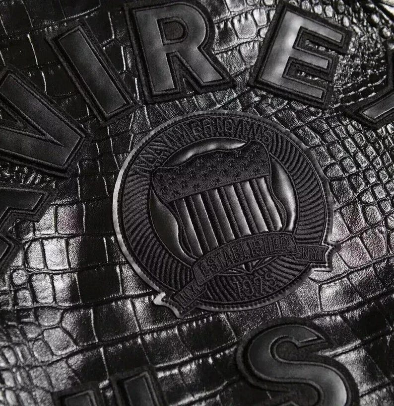 May include: A black material with a crocodile skin texture featuring large black stitched lettering and a circular emblem. The emblem displays a shield with stars and stripes, the text AMERICANS, ESTABLISHED, 1979, and NY in a circular border.
