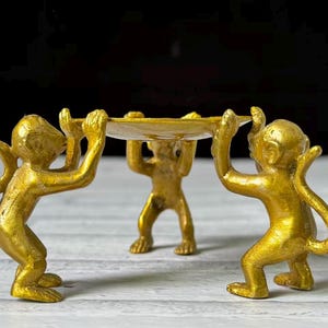 May include: A gold-colored decorative tray held by three monkey figurines. The monkeys are in a standing position, supporting a round, shallow tray. The background is dark, and the tray is on a white surface.