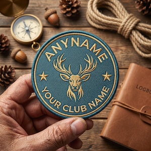 Könnte beinhalten: Rundes, teal-farbenes und goldenes gesticktes Abzeichen mit einem Hirschkopf und dem Text "ANYNAME YOUR CLUB NAME". Das Abzeichen wird in einer Hand gehalten, mit Kompass, Seil und Notizbuch im Hintergrund.