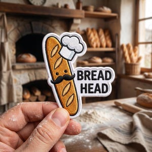 May include: Embroidered patch depicting a cartoon baguette wearing a chef's hat and a mustache, with the text "BREAD HEAD". The background shows a bakery with various loaves of bread.