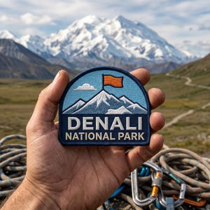 May include: A semi-circular embroidered patch featuring a light blue sky, a white mountain peak, and an orange flag. The text DENALI NATIONAL PARK is stitched in white and cream lettering on a dark blue background with a dark blue border.