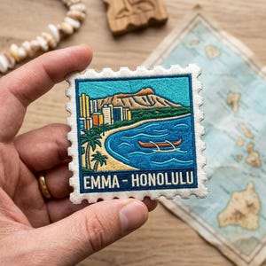 May include: Embroidered patch in the shape of a postage stamp depicting a Honolulu scene. The design includes a mountain, ocean, palm trees, buildings, and a canoe. The text "EMMA - HONOLULU" is at the bottom.