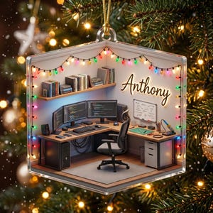 May include: A clear, hexagonal acrylic ornament featuring a miniature 3D office scene with a desk, computer monitors, a chair, and shelves. The name Anthony is written on the wall in script. Multi-colored string lights frame the interior scene.