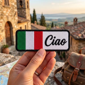 Puede incluir: Un parche rectangular con la bandera italiana y la palabra "Ciao" en escritura negra. El parche tiene un borde negro. El fondo muestra una ciudad europea con edificios de piedra y una vista panorámica.