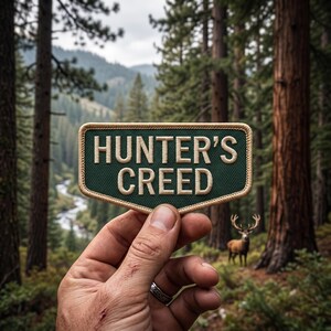 Könnte beinhalten: Ein rechteckiger gestickter Aufnäher mit dunkelgrünem Hintergrund und beigem Rand. Der Text HUNTER'S CREED ist in beigen Großbuchstaben in der Mitte des Aufnähers eingestickt. Der Aufnäher hat eine leicht zulaufende Unterkante.