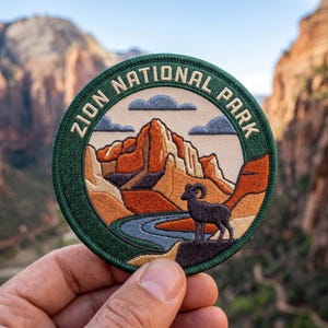 May include: Circular embroidered patch with the text "ZION NATIONAL PARK" in white on a green border. The patch shows a mountain landscape with a river, a bighorn sheep, and clouds. The colours include green, tan, orange, blue, and black.