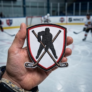 Könnte beinhalten: Ein Hockey-Patch mit einer schwarzen Spieler-Silhouette und gekreuzten Hockeyschlägern auf einem weißen Schild. Es hat einen roten Rand und eine schwarze Umrandung. Der Patch wird in einer Hand gehalten, mit einer Hockey-Eisbahn und Spielern im Hintergrund.